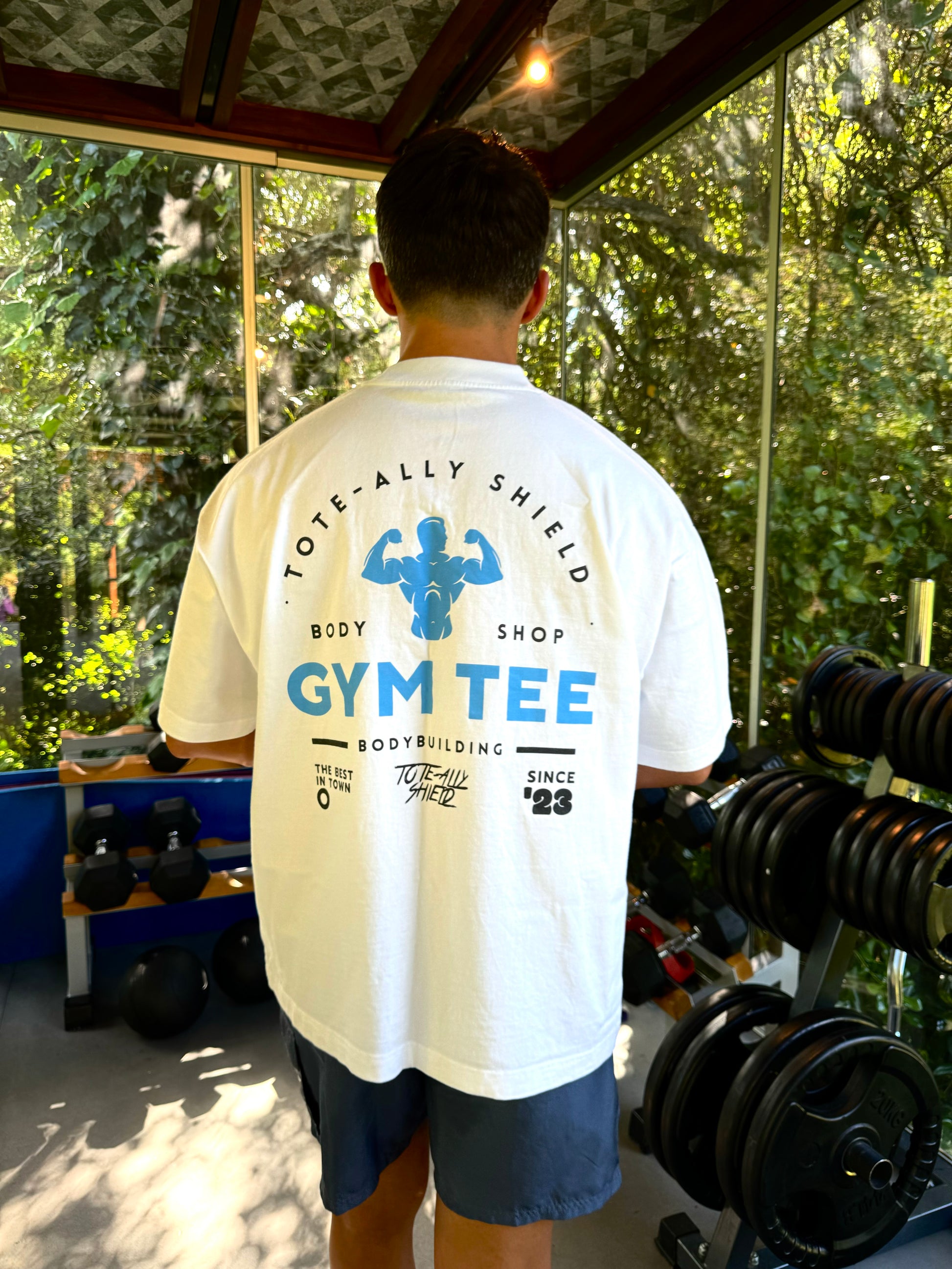 The back view of our white shortsleeved, heavyweight, oversized GYM T-shirt from TOTE-ALLY SHIELD GYM collection. Features bold GYM TEE logo emblazoned on center back. Made from heavyweight, 350GSM, 100% cotton for unparalleled quality and durability.