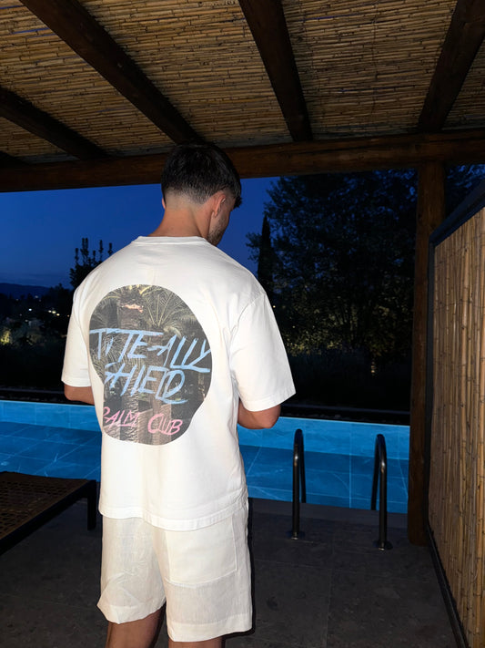 Back view of white short-sleeved, boxy-fit, heavyweight T-shirt featuring a large center-back graphic print. The ‘PALM CLUB’ design showcases a summer palm tree escape in a circular shape, with baby blue text highlighting the ‘TOTE-ALLY SHIELD’ logo and hot pink text highlighting 'PALM CLUB'. Picture Background poolside under the sun with a bamboo backdrop. 
