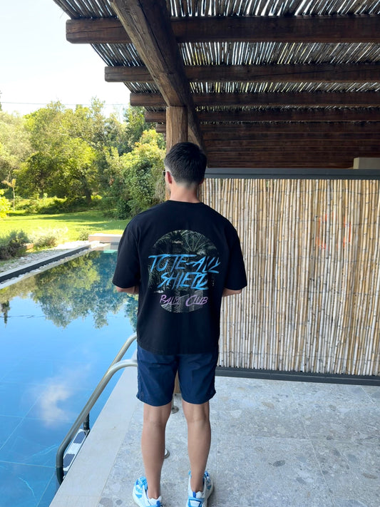 Back view of black short-sleeved, boxy-fit, heavyweight T-shirt featuring a large center-back graphic print. The ‘PALM CLUB’ design showcases a summer palm tree escape in a circular shape, with baby blue text highlighting the ‘TOTE-ALLY SHIELD’ logo and hot pink text highlighting 'PALM CLUB'. Picture Background poolside under the sun with a bamboo backdrop. 