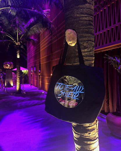 Front view of the TOTE-ALLY SHIELD Tote Bag in black canvas, featuring the PALM CLUB print on both the front and rear sides. The Tote bag is hanging on a palm tree under pink and blue mood lighting at night. 