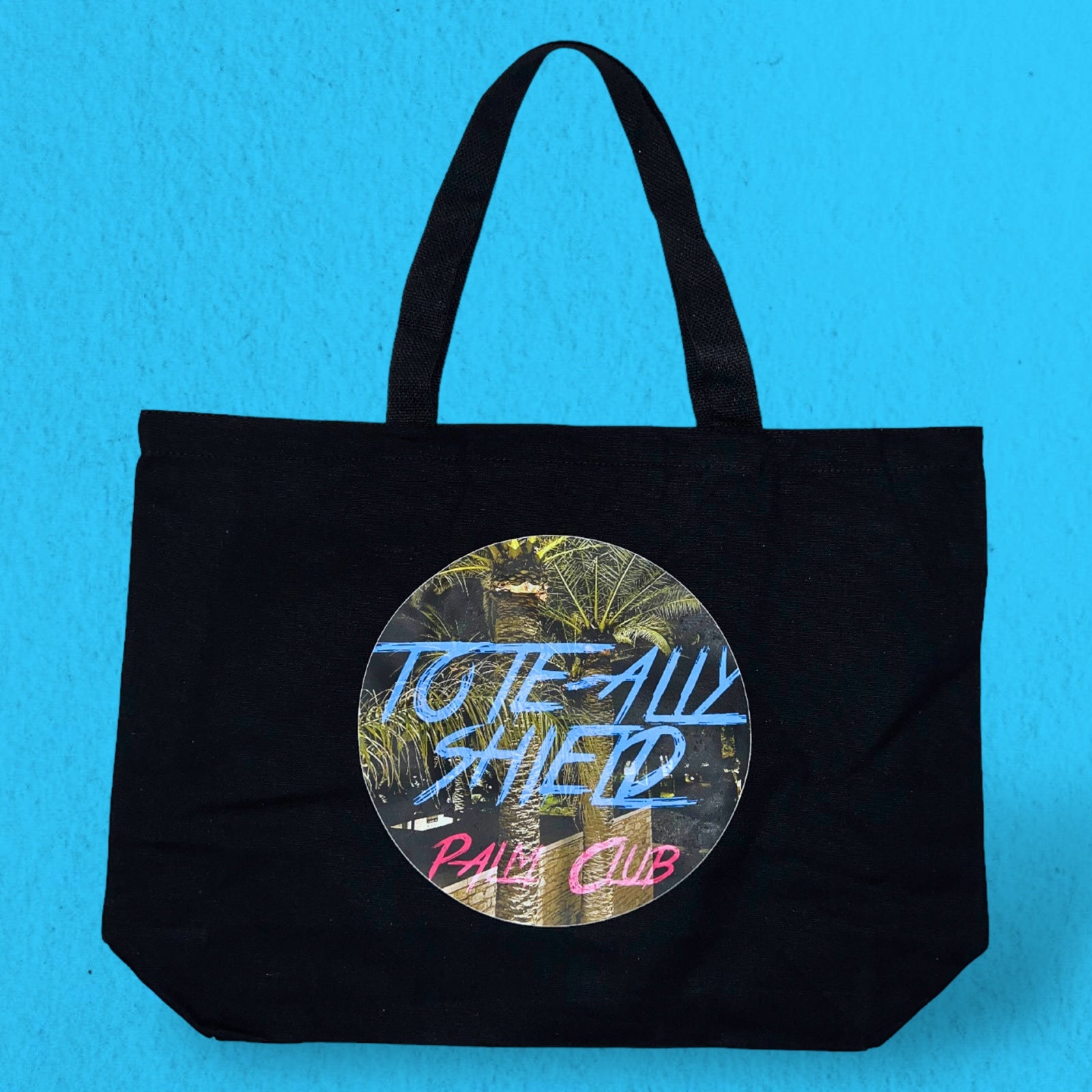 Front view of the TOTE-ALLY SHIELD Tote Bag in black canvas, featuring the PALM CLUB print on both the front and rear sides.